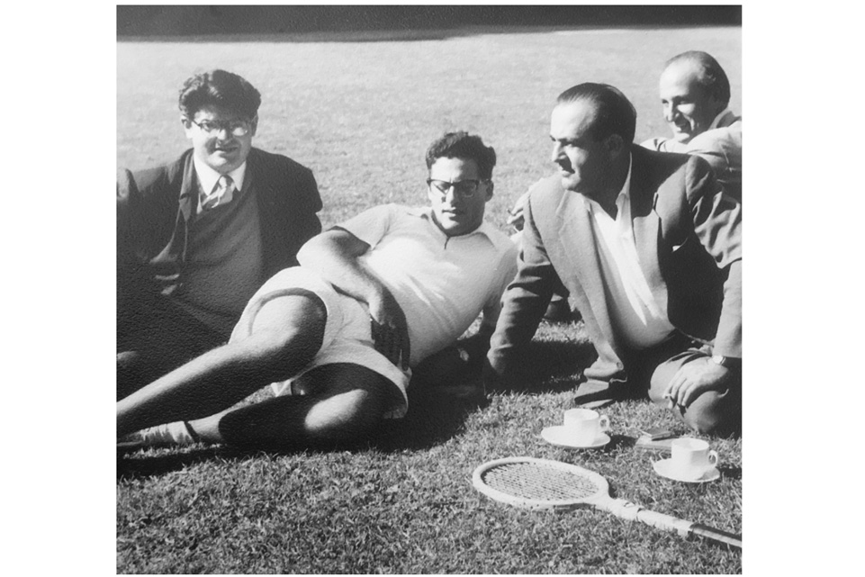 An old photo of a group of men, leaning on the grass, with a tennis racket and cups on the ground.